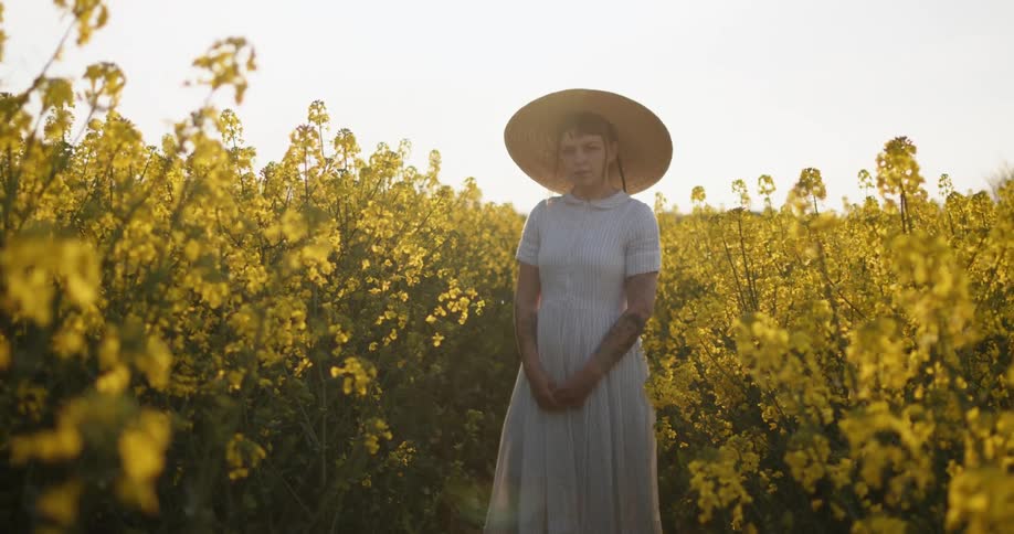 A woman in a dress and sun hat stands outside between rows of yellow flowering plants
