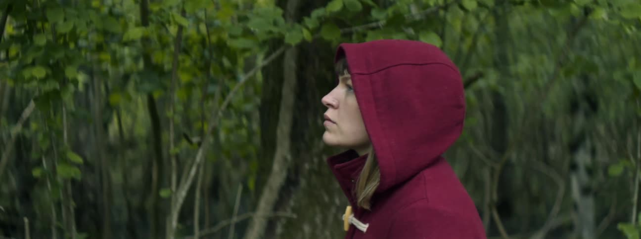 A woman in purple hooded jacket walks alone in a wooded area during the day