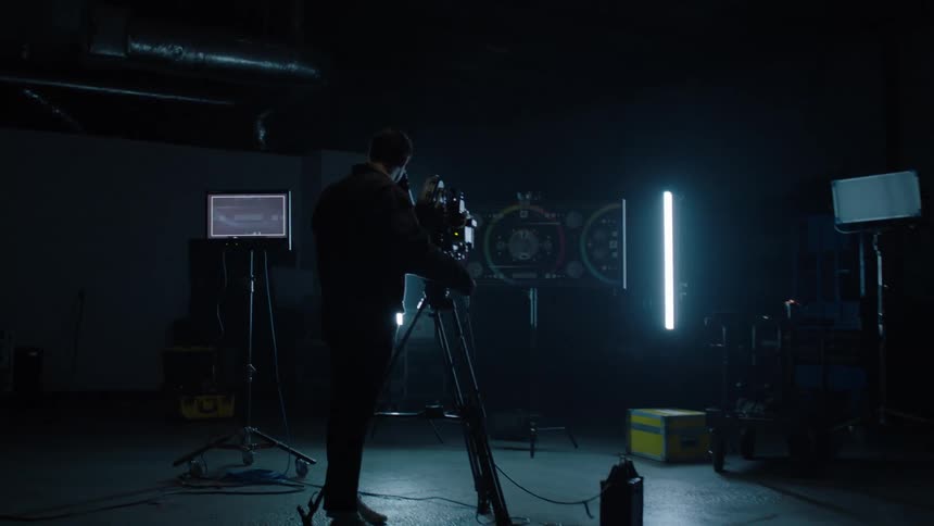A man stands behind a video camera on a tripod surrounded by screens and monitor and glowing lights in a dark room