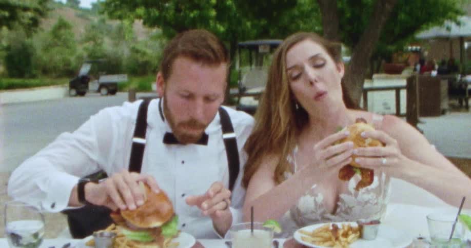 A bride and groom laugh as they eat burgers on their wedding day