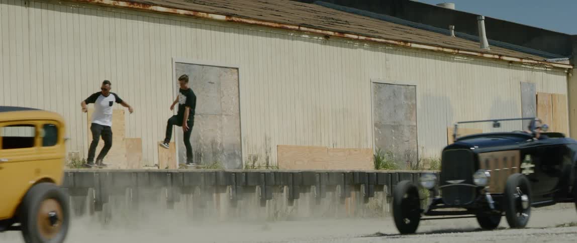 Two men skateboard on a raised area while classic cars drive past an abandoned building