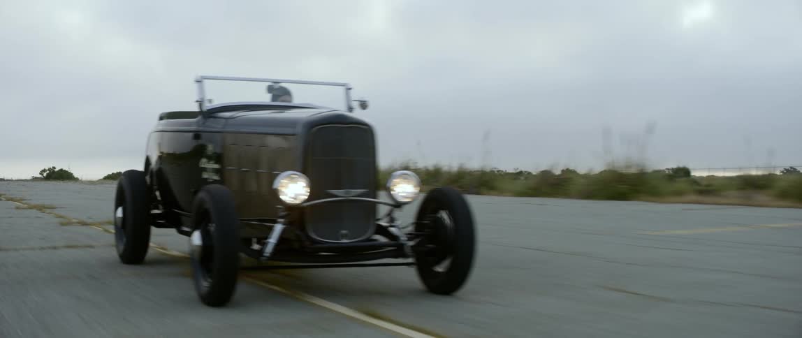 Two antique cars race on a paved road in the countryside under a cloudy sky