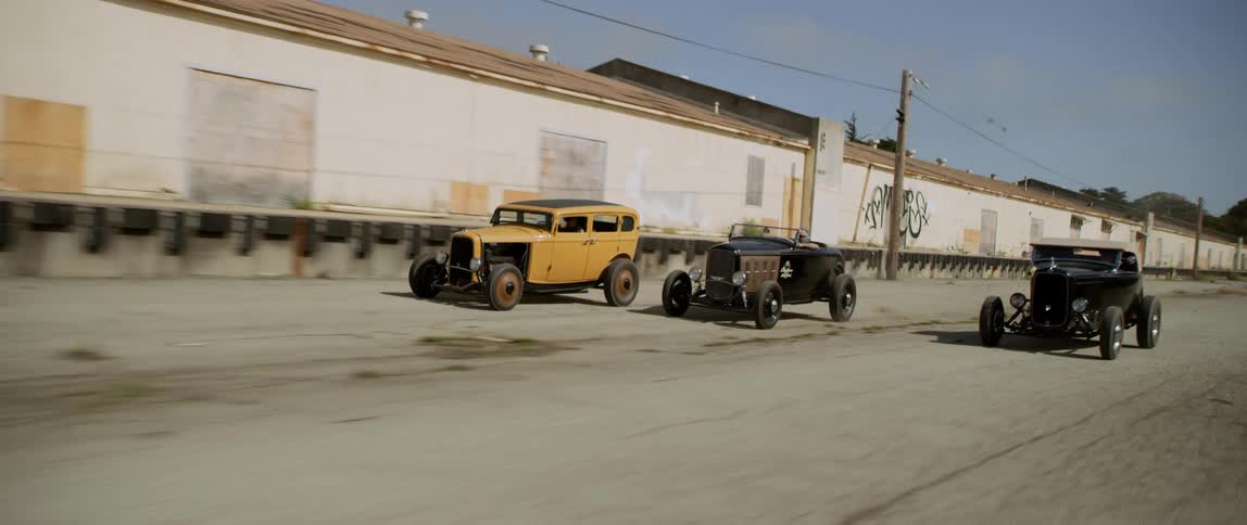Three vintage cars race on an empty strip near a long boarded up building on a sunny day