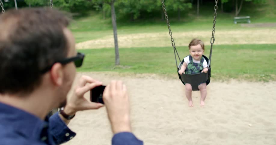 A father takes a photo with his phone of his son in a baby swing at a park
