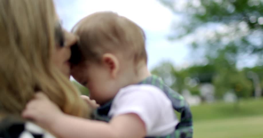 A woman with sunglasses holding a baby boy and kisses him on the forehead