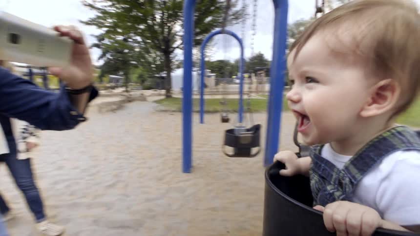 A dad holds up his phone and films his smiling laughing child on a swing