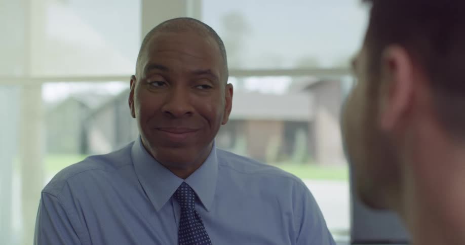 Black man and white man talk in a room as cars go by outside