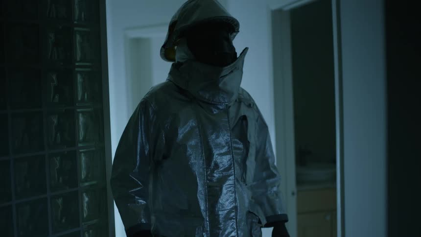 A person in full body reflective personal protective equipment walks through a hallway illuminated only by a rapidly flashing pale blue light