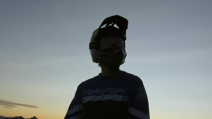 A person in a motorcycle helmet standing outdoors as the low sun shines through the peaks of a far off mountain
