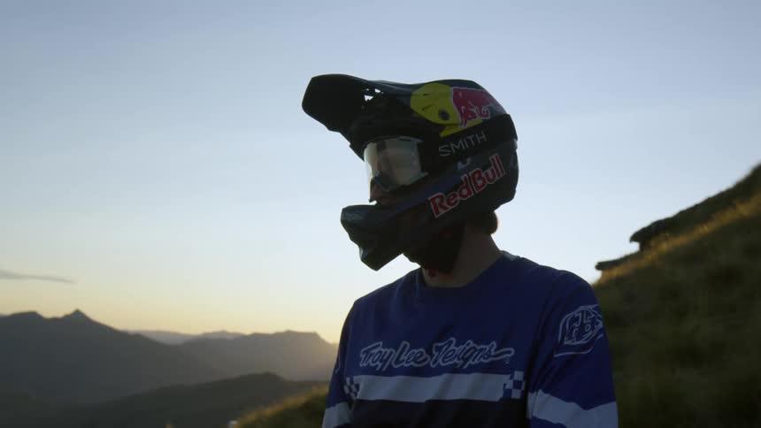 Man wearing a motorcycle helmet with the visor up sits with mountains in the distance