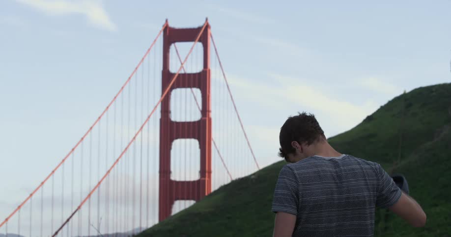 Man removes his hat as he stands near a hill and a bridge