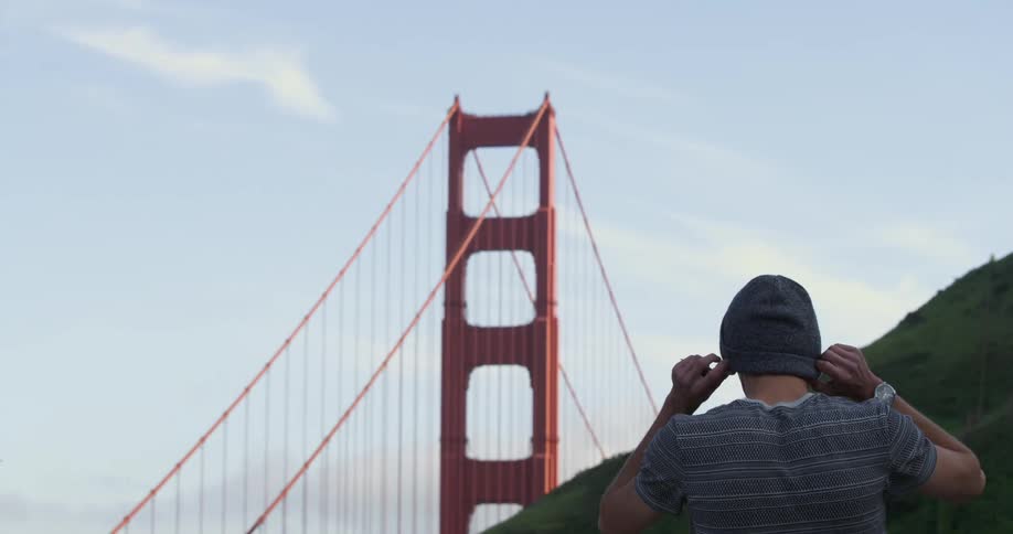 A man puts on a beanie and walks away with the golden gate bridge in the background