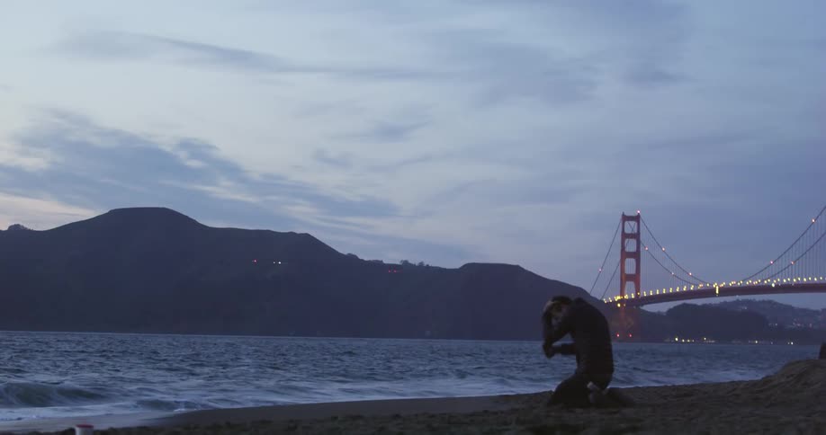 A man walking on the shore towards the water drops to his knees and hits the sand with his fist and puts his head in to his hands and cries
