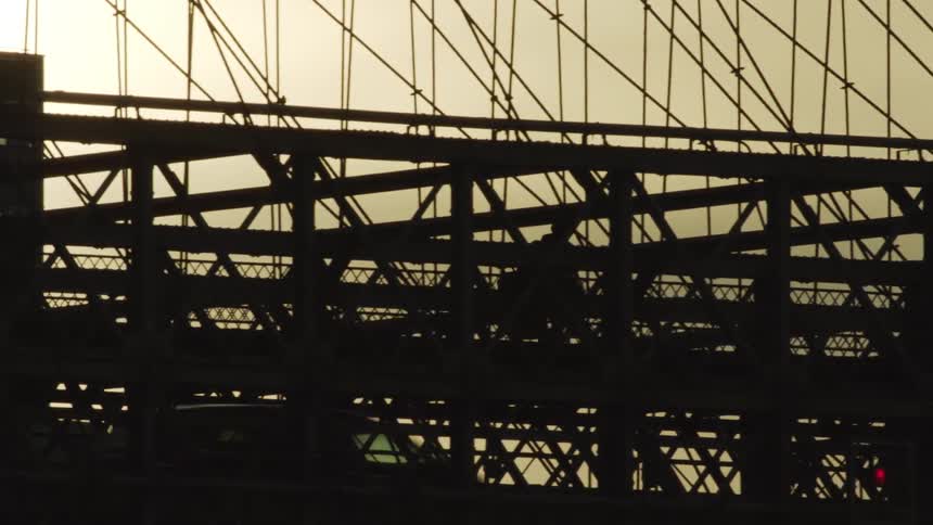 People walk above Brooklyn Bridge in New York at Sunset with traffic moving by on a metal bridge