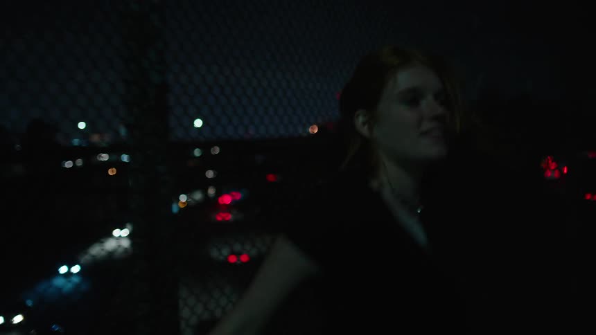 Woman with sparkling pendant walks happily and touches wire fences as she crosses a overpass at night