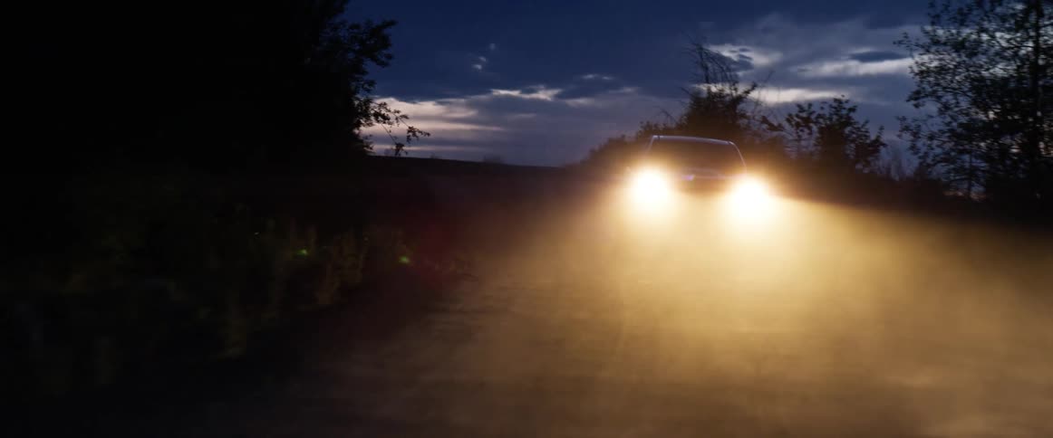 Car headlights glare in fog as car drives along road between trees at night