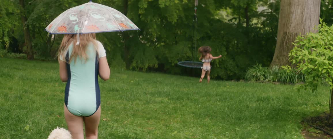 A little girl runs to a swing outside in the rain while her sister carries an umbrella follows her with their dog