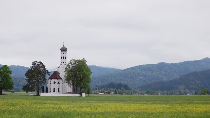 A white building with a tall tower and dome sits in the middle of a field near hills