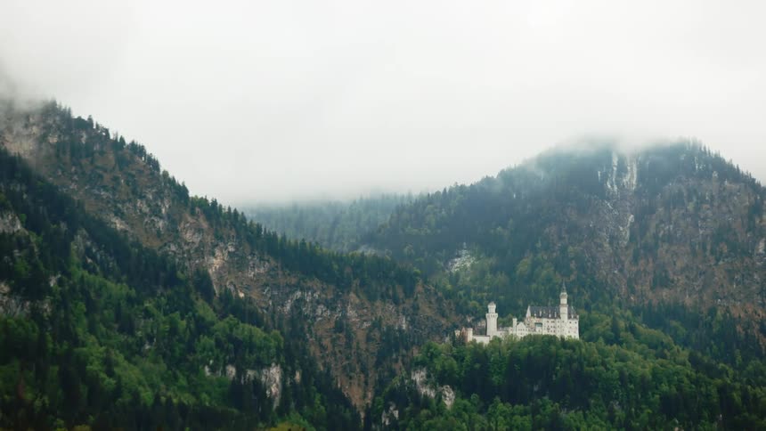 A large building on a verdant mountain between two peaks that are shrouded in clouds
