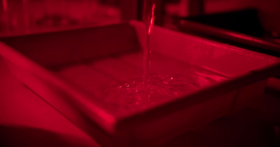 A person pouring liquid from a beaker into a plastic ban in a dark room illuminated by a red light