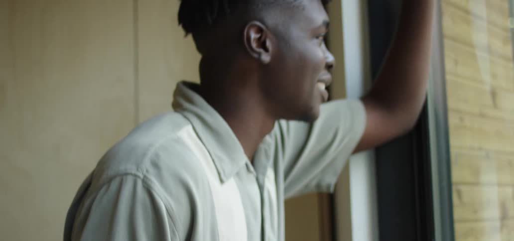 A man smiles as he looks out the window of his apartment while also talking on his phone