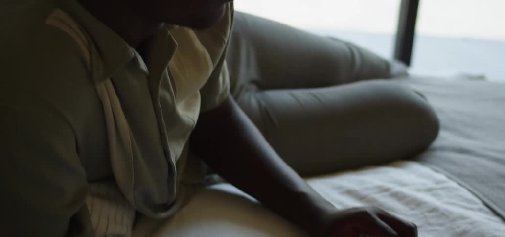Black man with grey polo shirt and pants lies down on a bed and types on a laptop