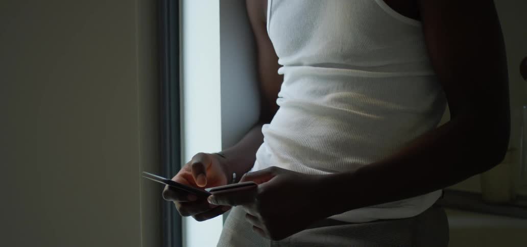A young black man leans against the wall next to the window while using his phone to make a purchase