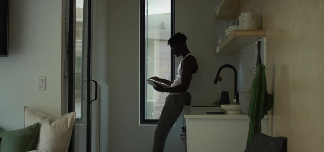Black man with a white undershirt leans against a kitchen counter and turns the pages of a book