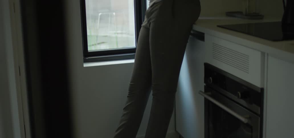 Black man with a white undershirt and grey pants uses a smartphone standing in the kitchen by a window