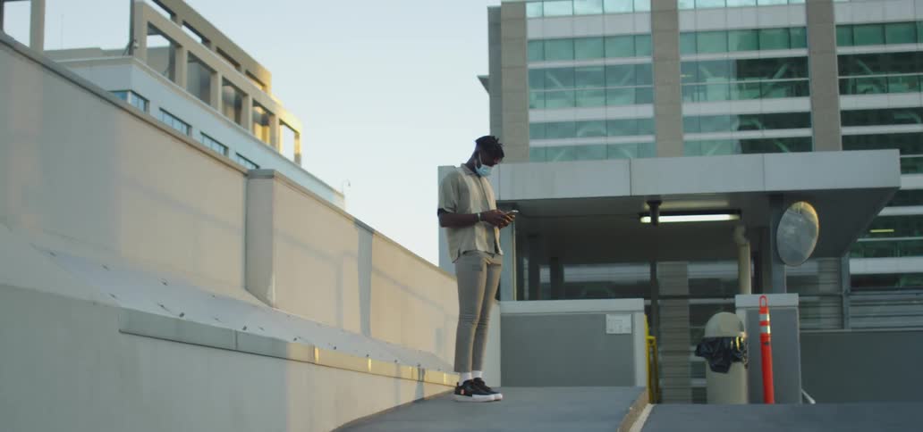 Black man with a grey polo shirt and a blue face mask stands and uses a smartphone