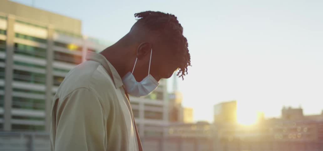 A black teenager in a face mask stands on a roof top and texts