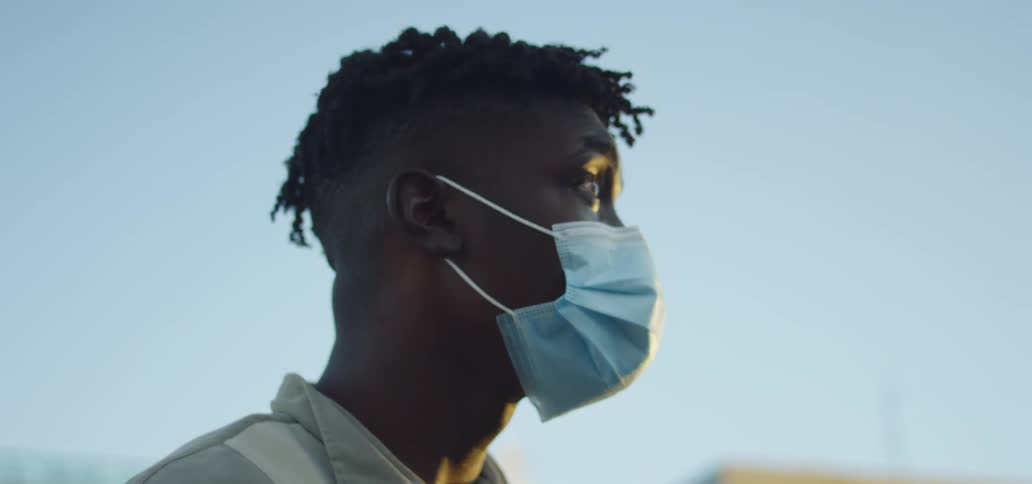 Black man with grey polo shirt and a blue face mask uses a smartphone as the sun goes down