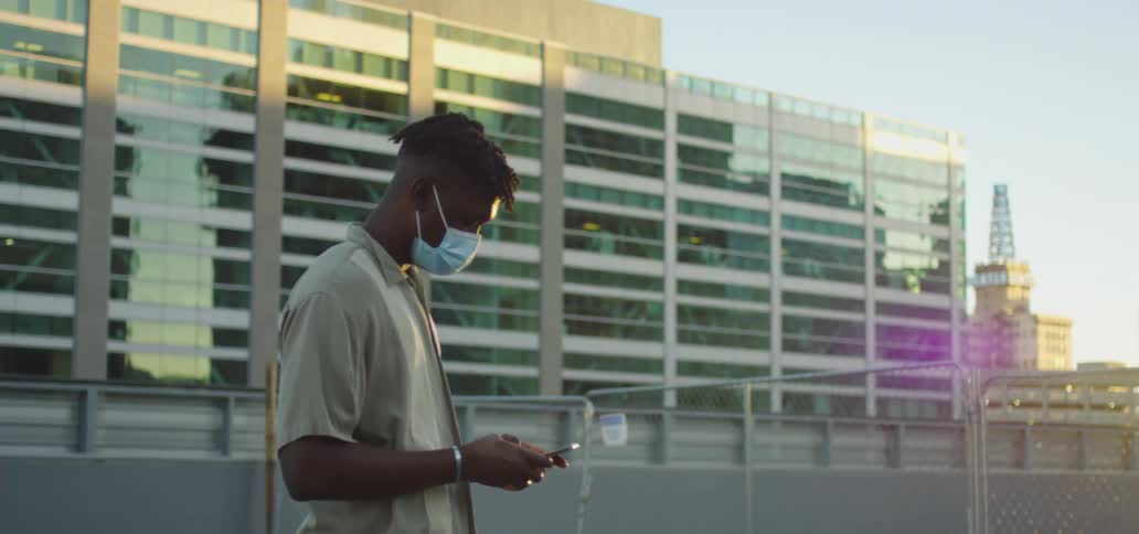 Young man wearing face mask types on his phone while standing on a roof at sunset