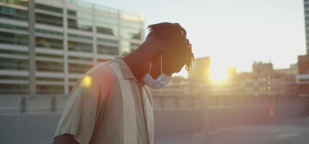 A man wearing a disposable protective mask stands in a parking lot looking around at the building and then back down