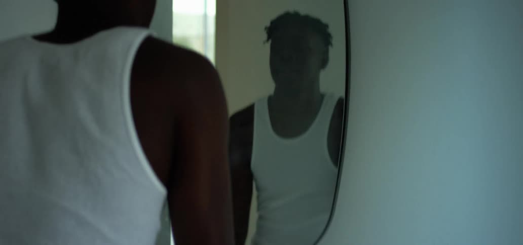 Black man in a white sleeveless shirt looks in an oval mirror