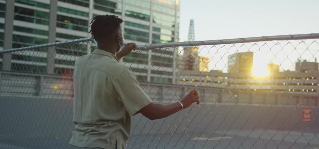 A frustrated black teenager in a face mask stands behind a chain link fence in the city