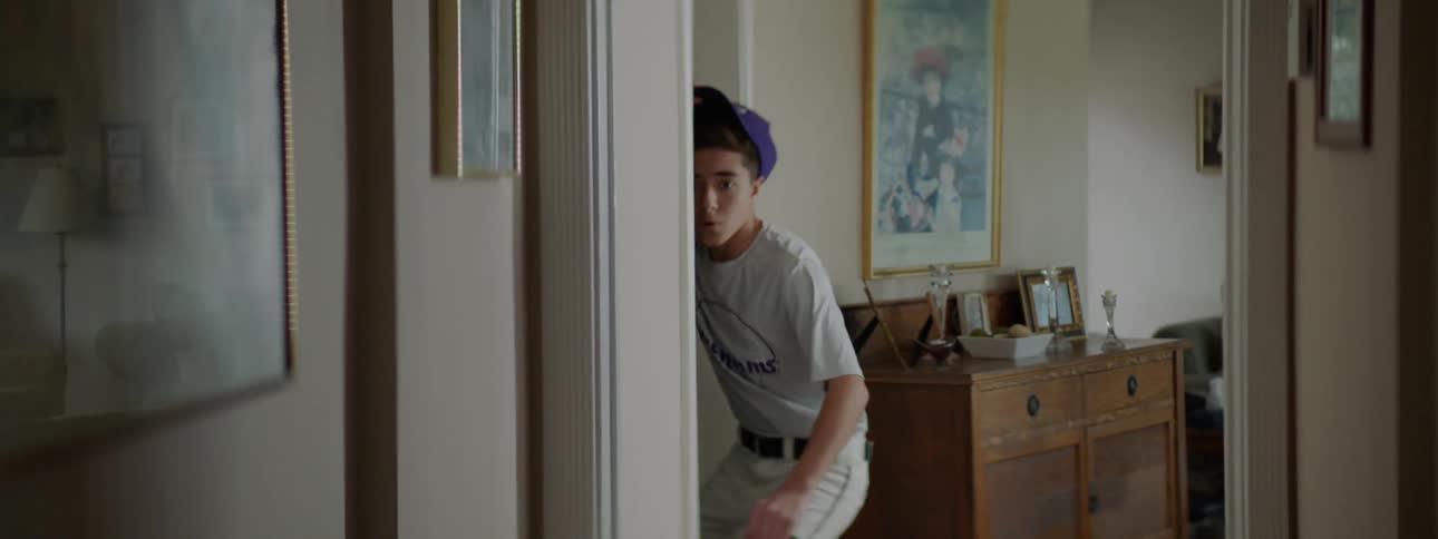 A boy in a baseball uniform is walking through his house and then runs in to another room