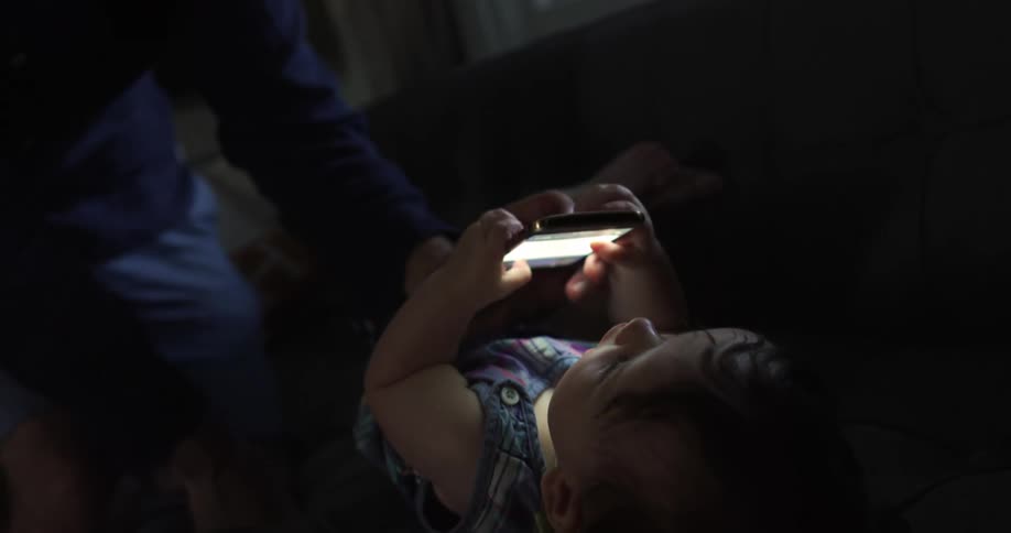 A baby holds a cell phone and stares it as an adult sits with them