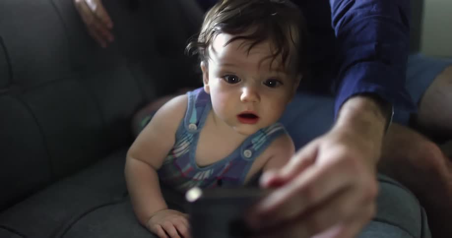 Baby girl looks at smartphone screen in surprise while lying on a couch