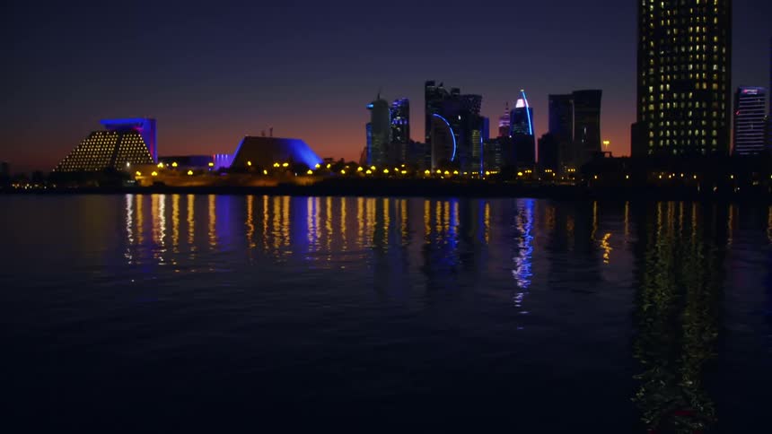 Buildings along a city skyline are lit with blue lights that reflect in the ocean before it