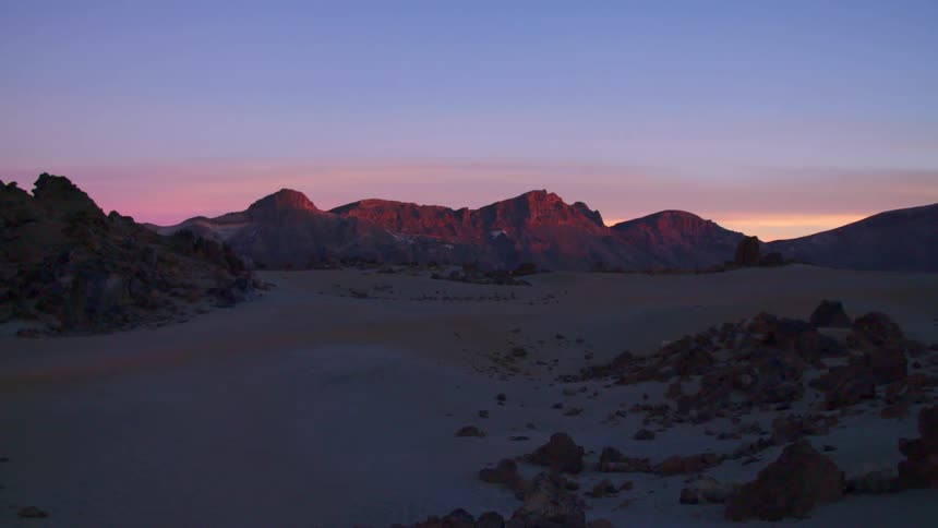 A desert with rocks sits near a mountain range at sunrise