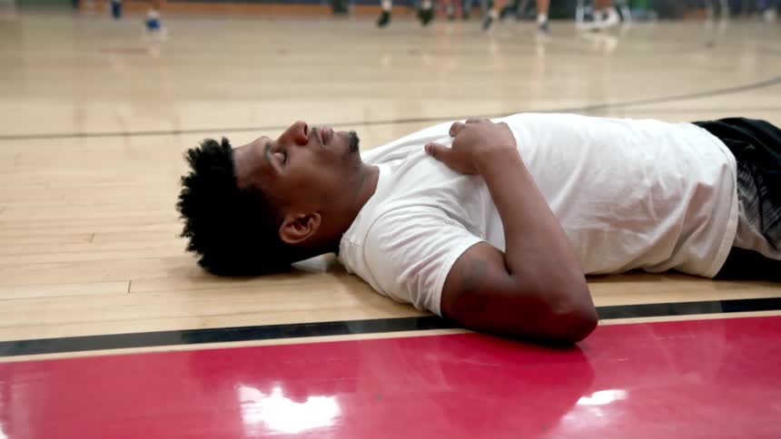 A man resting on the floor of a basketball court as other people play basketball in the background