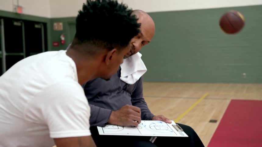 A teenager dribbles a basketball while another sits and listens to his coach and looks at his clipboard