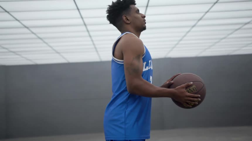 Black man in a blue jersey stands in a gym holding a basketball