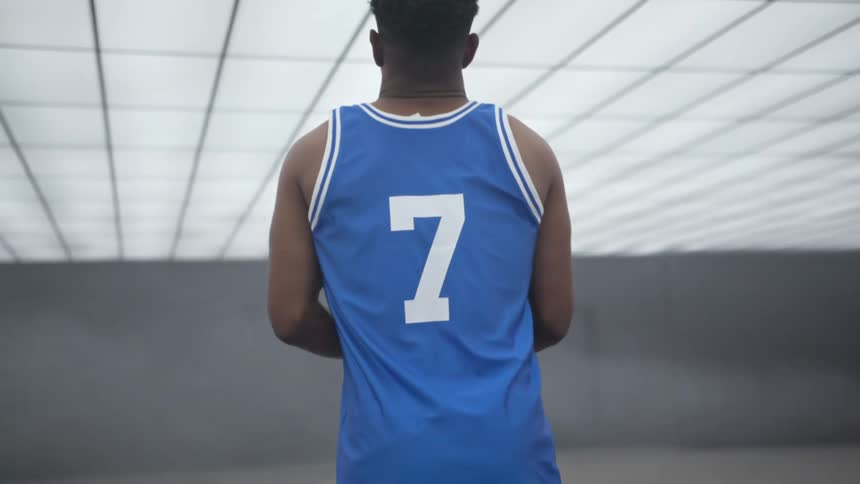 A black man in a uniform and holding a basketball stands in a room with a ceiling made out of lights