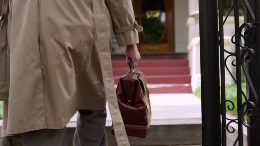Man with a trench coat and pants holds a brown bag and walks up to a door
