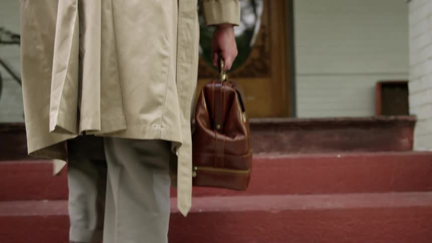 A person wearing a trench coat and carrying a leather bag ascends a set of red stairs towards the front door of a house