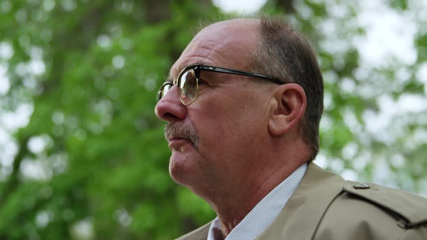 A man with glasses shirt and coat looking up with trees and the sky in the background