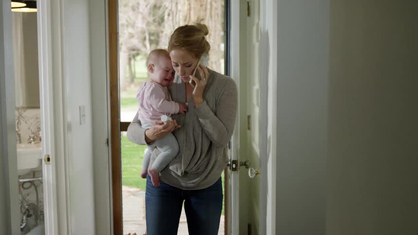 A mother holds her crying baby as she looks at a label on some medication and makes a phonecall