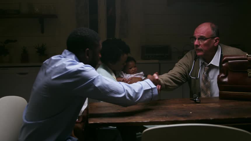 A doctor shakes hands with a black man while sitting at a table with the mans family